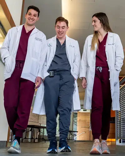 Three medical students in white coats walk together through hallway during rounds