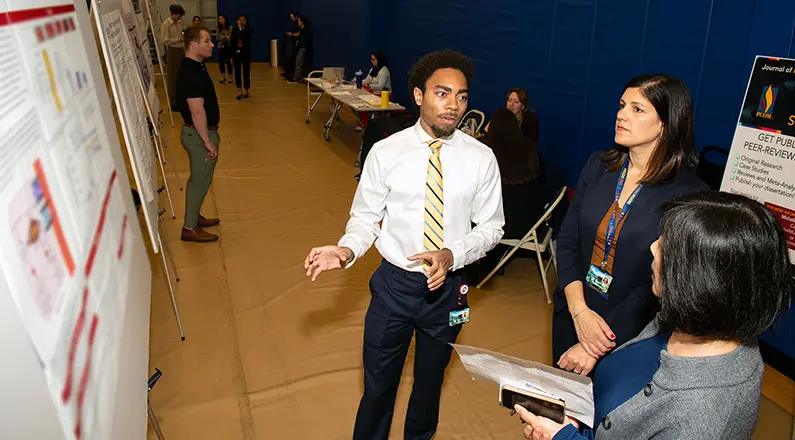 PCOM grad student Cameron Stinson (MS/Biomed '24, DO '28) talks to faculty in front of his research poster.