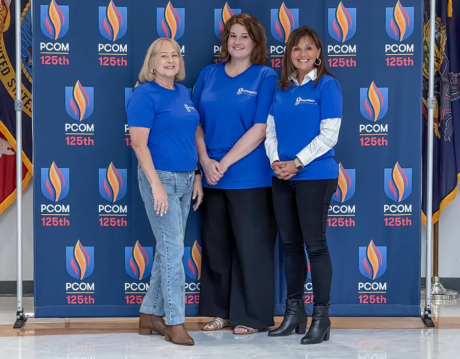 Three members of the Pharmacy Experiential Education team smile in front of a PCOM logo backdrop